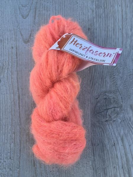 Ein fluffiger, handgefärbter Strang Wolle in leuchtendem Korallorange mit feinen rosa und apricotfarbenen Nuancen. Der Strang liegt auf hellem Holz. Das Garn wirkt weich, flauschig und leicht glänzend.